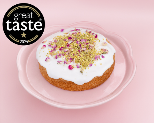 Decorative cake with white icing and rose petals on a pink background, featuring the Great Taste logo.